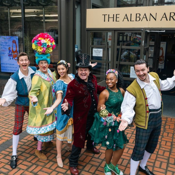 A group of six people in colourful, theatrical costumes stand happily with arms open in front of The Alban Arena. They are smiling and standing on a brick pavement near a flowerbed, seemingly promoting a performance or event at the venue.