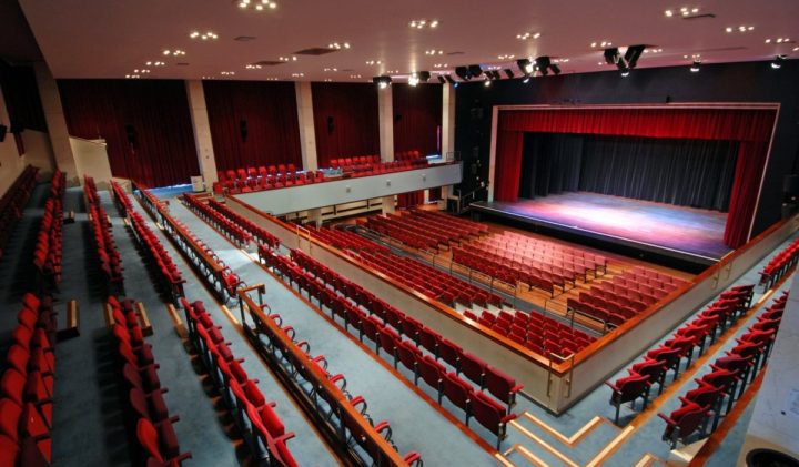 A view of a large, empty Alban Arena theatre with red seats arranged in multiple levels, facing a wide stage with a red curtain. The theatre features balcony seating, aisle pathways, and modern lighting fixtures illuminating the space.