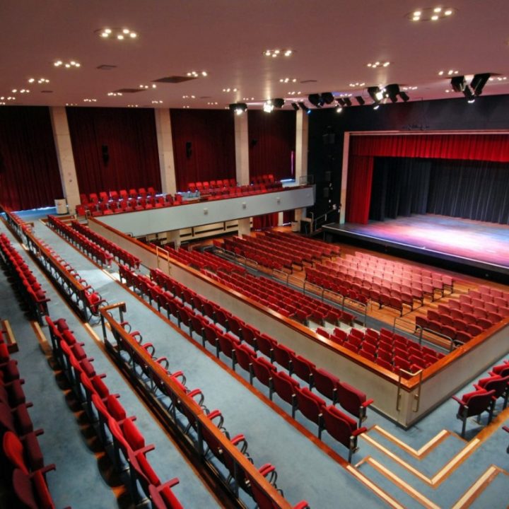 A view of a large, empty Alban Arena theatre with red seats arranged in multiple levels, facing a wide stage with a red curtain. The theatre features balcony seating, aisle pathways, and modern lighting fixtures illuminating the space.