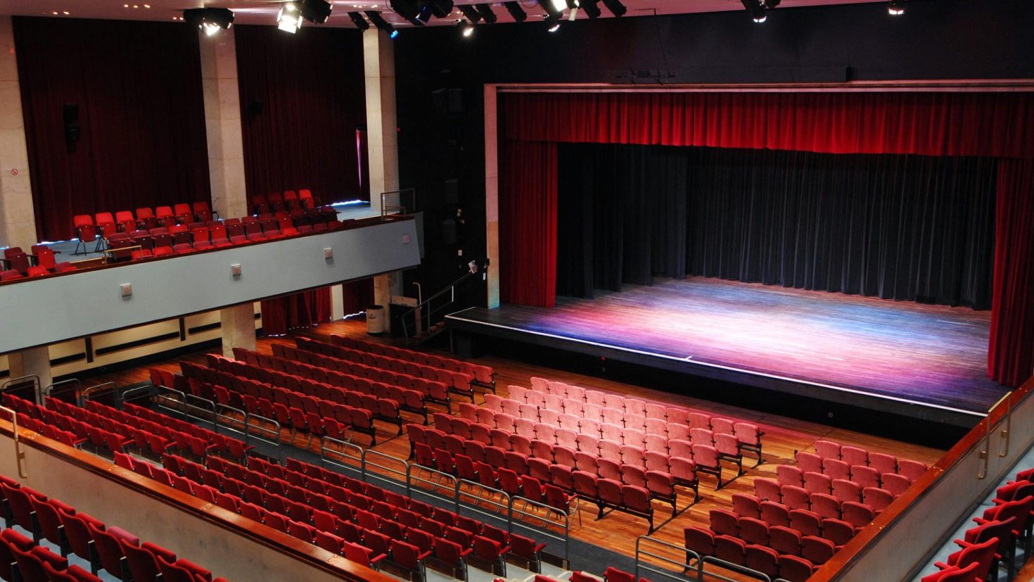 A large Alban Arena theatre with red cushioned seats facing a wooden stage with red and black curtains. The theatre has multiple seating levels, with a balcony and ground floor section. Stage lights hang from the ceiling, and the room is empty.