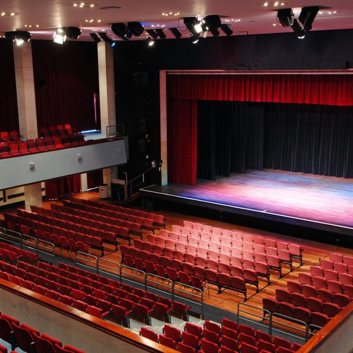 A large Alban Arena theatre with red cushioned seats facing a wooden stage with red and black curtains. The theatre has multiple seating levels, with a balcony and ground floor section. Stage lights hang from the ceiling, and the room is empty.