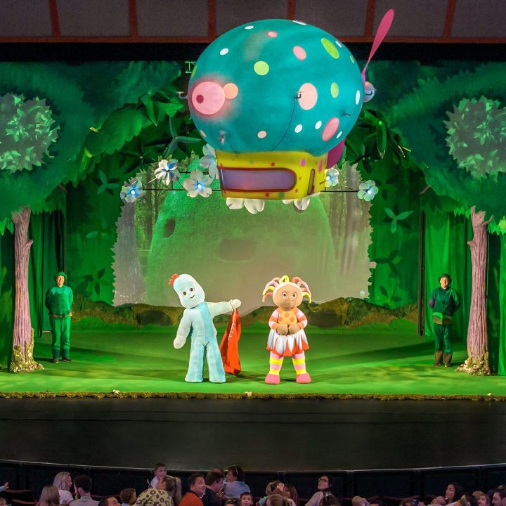 Stage production of the children’s show “In the Night Garden” featuring characters Igglepiggle and Upsy Daisy. The set is a vibrant, whimsical forest with green trees, and a large colourful hot air balloon suspended above. An audience is visible in the foreground.