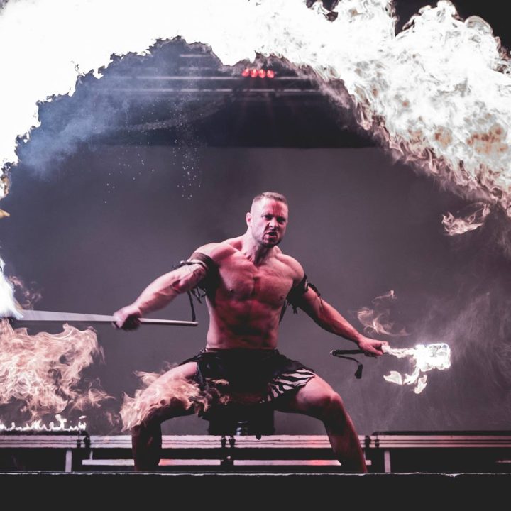 A muscular man performing a fire dance stands in a low stance holding a flaming staff with flames creating an arch above him. He is shirtless, wearing black shorts, and is on a dimly lit stage with fire effects in the background.