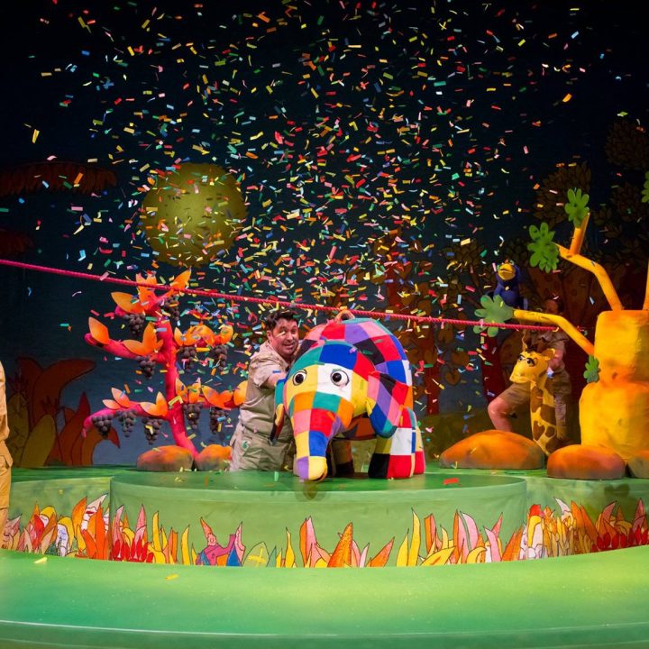 A colourful stage performance featuring actors with puppets, including elephants and a patchwork-patterned elephant, surrounded by vibrant trees and plants. Confetti rains down, enhancing the festive atmosphere. The backdrop is dark with brightly coloured decorations.