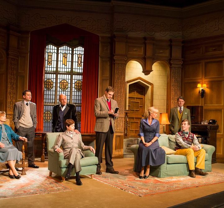 A group of actors dressed in period costumes are on a stage set resembling an ornate room with wooden panelling and a large stained glass window. They are positioned around two armchairs and a sofa, engaged in a scene, with one person standing and reading aloud.