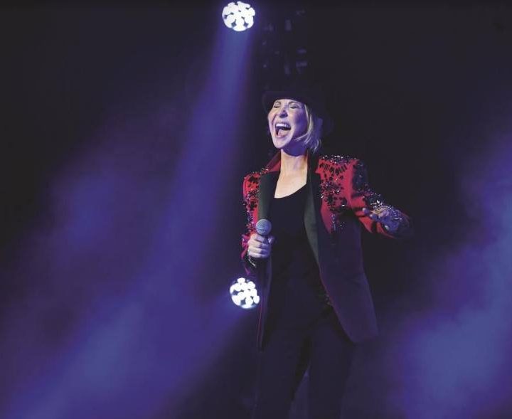 A person wearing a red and black sequined jacket and a black hat is performing on stage, holding a microphone and singing passionately. The background is dark with purple and blue stage lights.