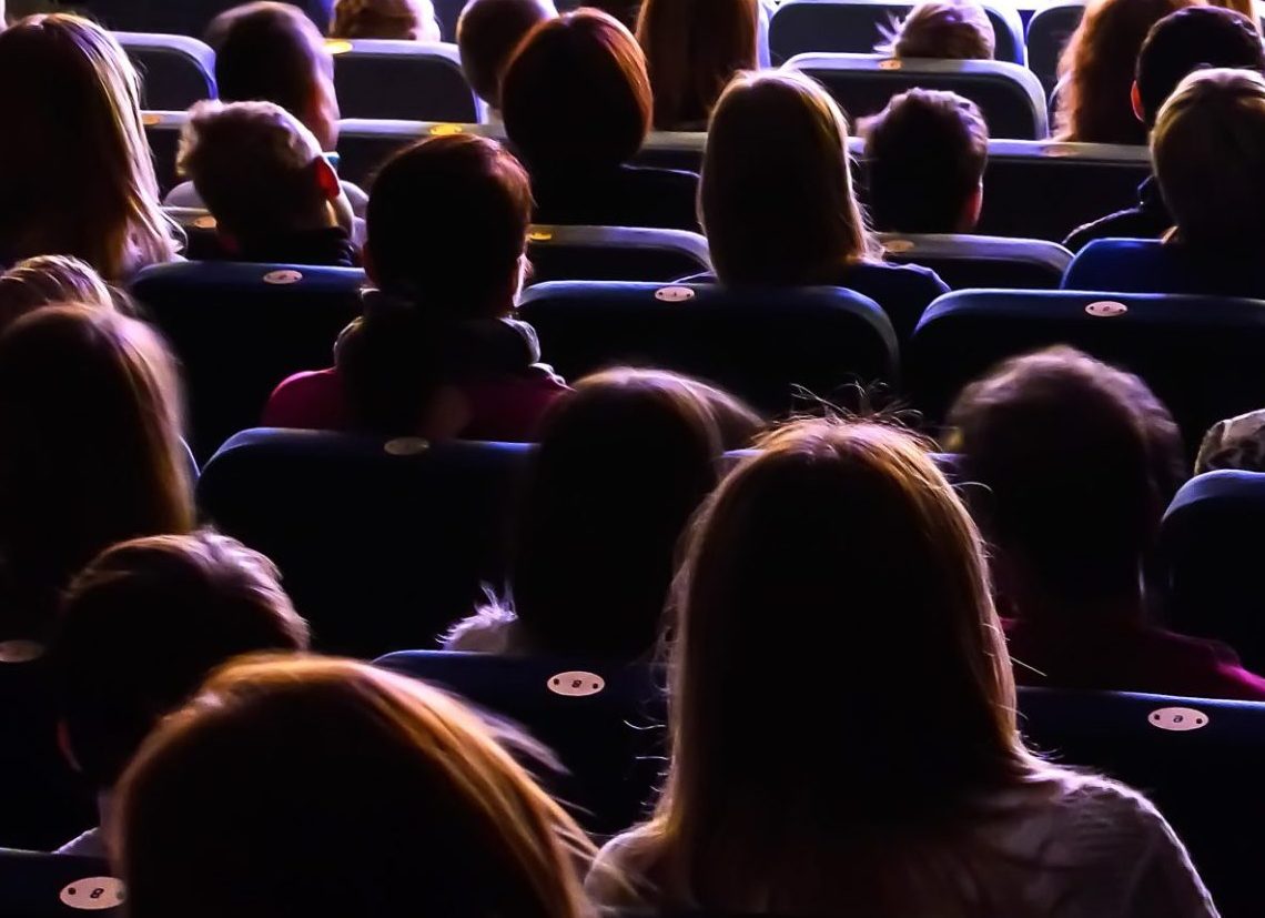 A group of people are seated in a theatre, facing forward towards the screen or stage. The audience is seen from behind, with dim lighting creating silhouettes and shadows, emphasising their heads and shoulders.