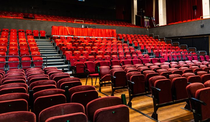 An empty theatre with rows of red seats and a black stage. The seats are arranged in an ascending manner, with a few tables covered in red cloth positioned in the centre of the highest row. The walls and ceiling are dark, with overhead lights illuminating the space.