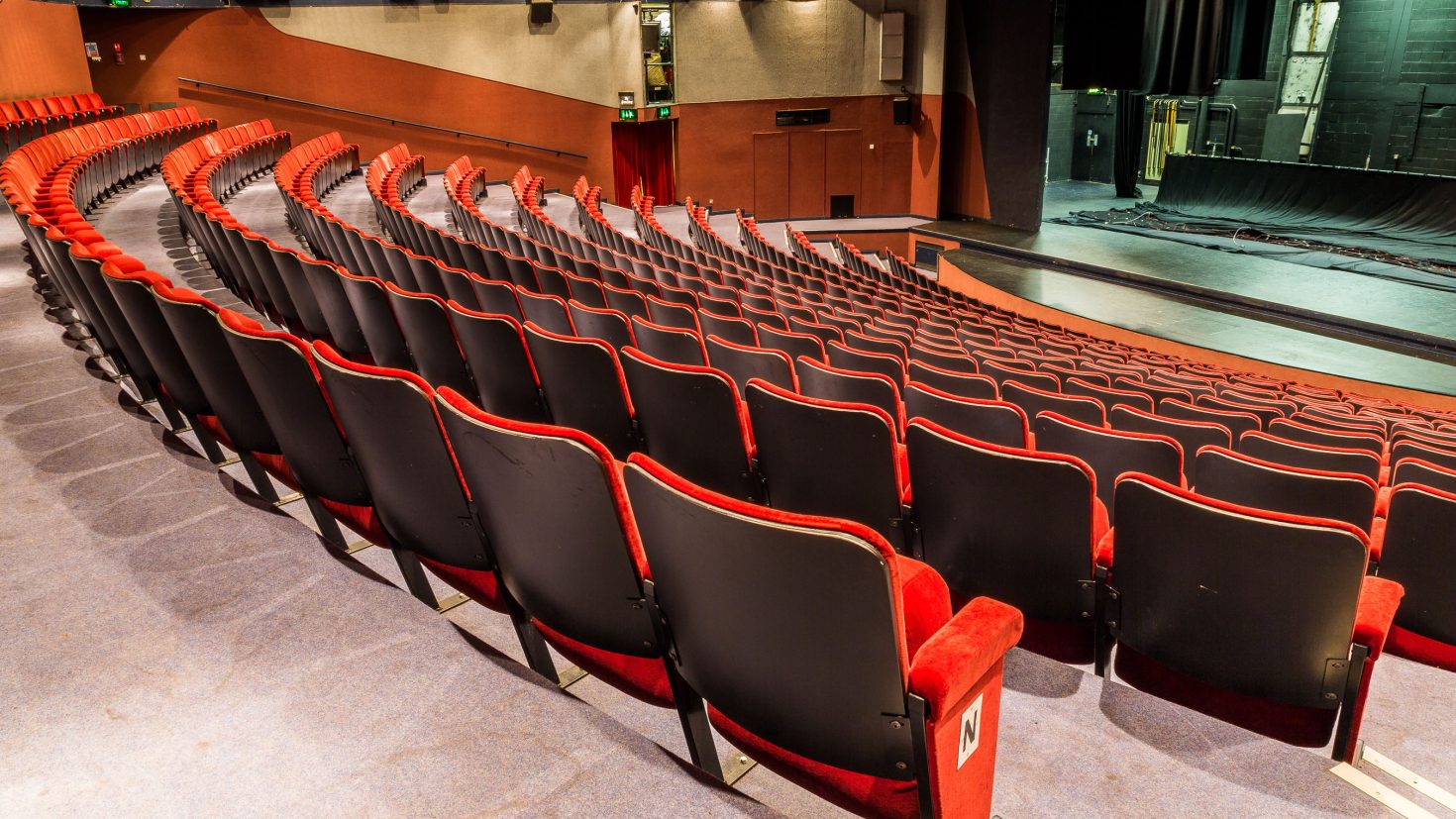 An empty, modern theatre with rows of black and red seats facing a stage with a closed black curtain. The theatre is well-lit, with a slightly curved seating arrangement. The stage area is partially visible, showing some backstage equipment.