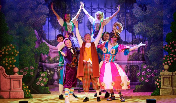 A group of performers stand on a brightly lit stage in colourful, eccentric costumes, including vibrant dresses, vests, and hats. They are posing energetically, some with arms raised, against a whimsical backdrop of a garden with flowers and foliage.