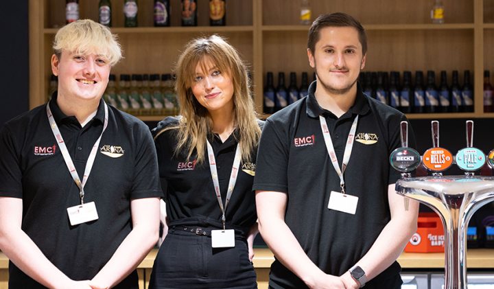 Three people in black uniforms stand smiling behind a bar. They wear name tags and the bar features four labelled beer taps. Bottles are neatly arranged on the shelves in the background.