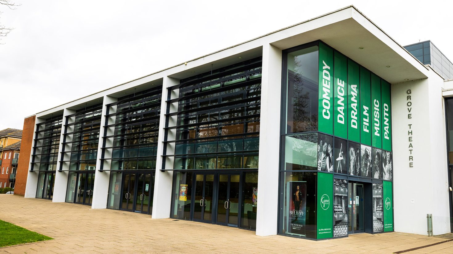 A modern building with a white façade and large glass windows. It features banners promoting comedy, dance, drama, film, and music. The side of the building reads "Grove Theatre". The entrance has multiple doors and the surroundings include brick pavement and nearby trees.