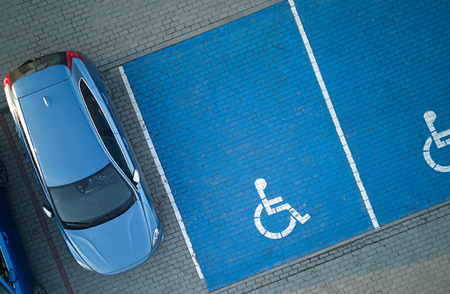 A blue car is parked partially over the line, encroaching into an empty disabled parking space marked with a wheelchair symbol. The asphalt is a mix of regular grey and blue pavers. Another disabled parking space is also visible and unoccupied.