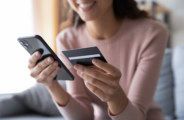 A person holding a smartphone in one hand and a credit card in the other, smiling. The focus is on the hands and the objects being held. The background is softly blurred.