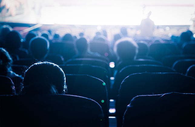 An audience sits in a dark theatre, facing a brightly lit stage or screen. The lighting casts the silhouettes of the seated people, creating a contrast between the dark seats and the illuminated area in front.