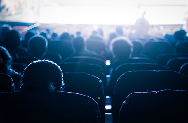 Audience seated in dimly lit Grove Theatre with a bright screen in the background.