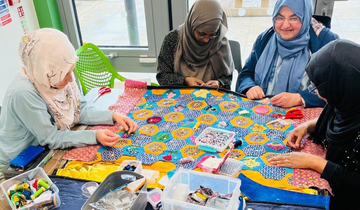 Four people sewing fabric for a creative newsletter at a table with craft supplies.