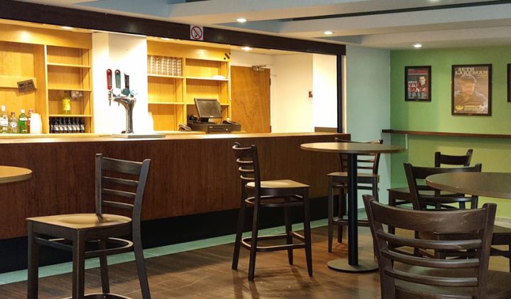 A modern, clean bar with a wooden counter and neatly arranged high chairs and tables. The area is well-lit with recessed ceiling lights. A selection of bottles and a beer tap are visible behind the counter. The walls are green with framed posters.