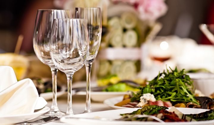 A close-up of a dining table set with empty wine glasses and plates of various salads and appetizers. A folded white napkin is on the left side, and blurred floral arrangements and additional glassware are in the background, creating an elegant atmosphere.