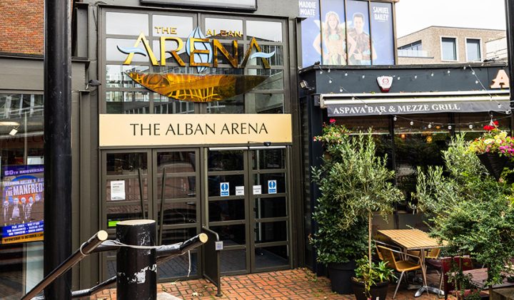 The front entrance of The Alban Arena, with its sign prominently displayed. Adjacent to the arena is Aspava Bar & Mezze Grill, featuring outdoor seating and decorative plants. Posters are visible on the arena's windows, and a brick path leads up to the entrance.