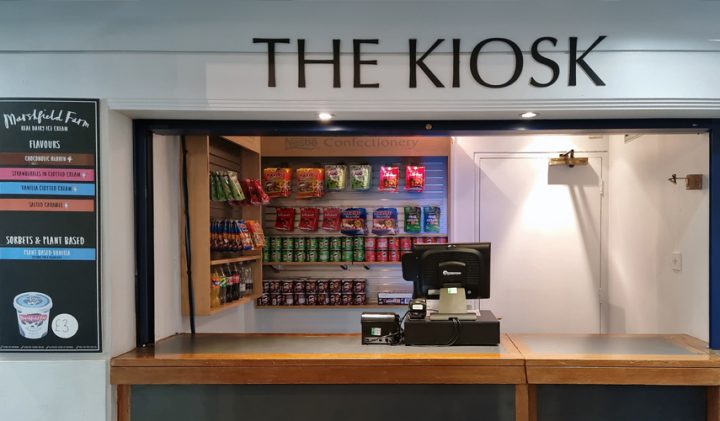 A small kiosk with a wooden counter and a sign overhead reading "THE KIOSK." Behind the counter is a shelf stocked with various snacks and confectioneries. A cash register is placed on the counter, and a menu board is displayed on the left side.