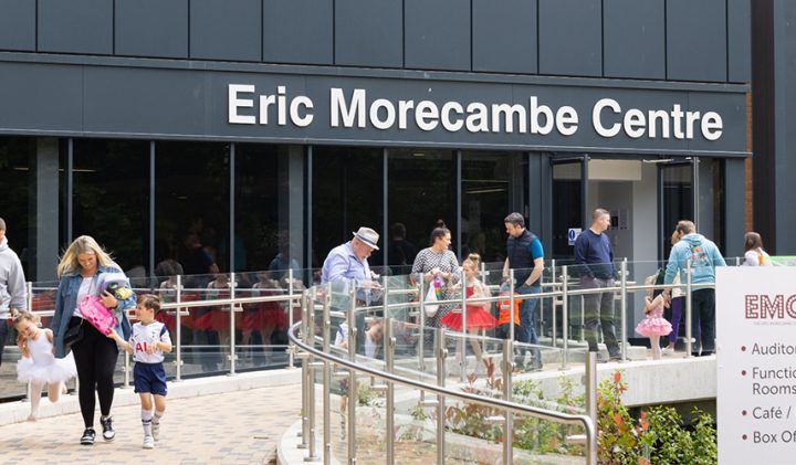 People walk up a gently sloping ramp towards the entrance of the Eric Morecambe Centre. Children in colourful outfits, including tutus, along with adults, are visible. A sign beside the ramp lists amenities such as an auditorium, function rooms, a café/bar, and a box office.