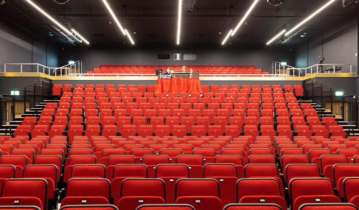 An empty theatre with rows of red seats and a black stage. The seats are arranged in an ascending manner, with a few tables covered in red cloth positioned in the centre of the highest row. The walls and ceiling are dark, with overhead lights illuminating the space.