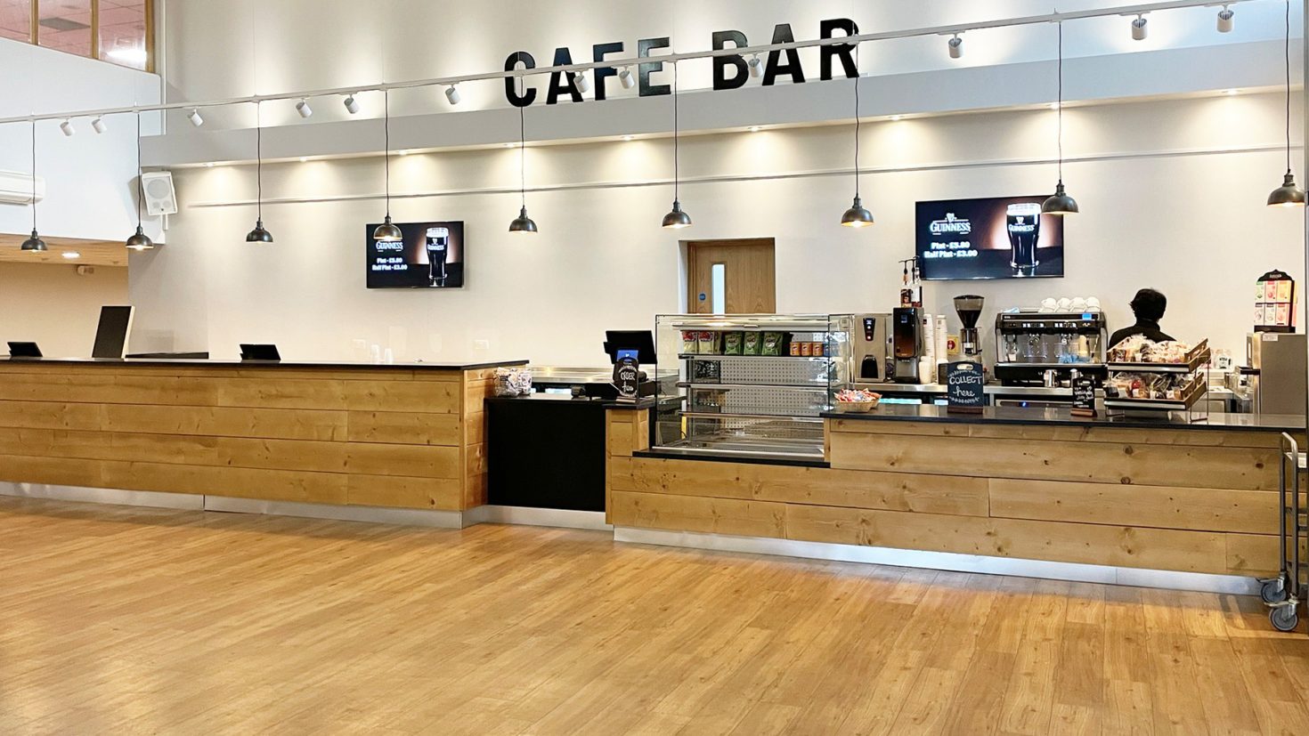 A modern café bar with wooden countertops and a clean, minimalist design. It features a large coffee machine, a display case with pastries, and digital menu screens. Pendant lights hang from the ceiling, and the floor is a light-coloured wood.