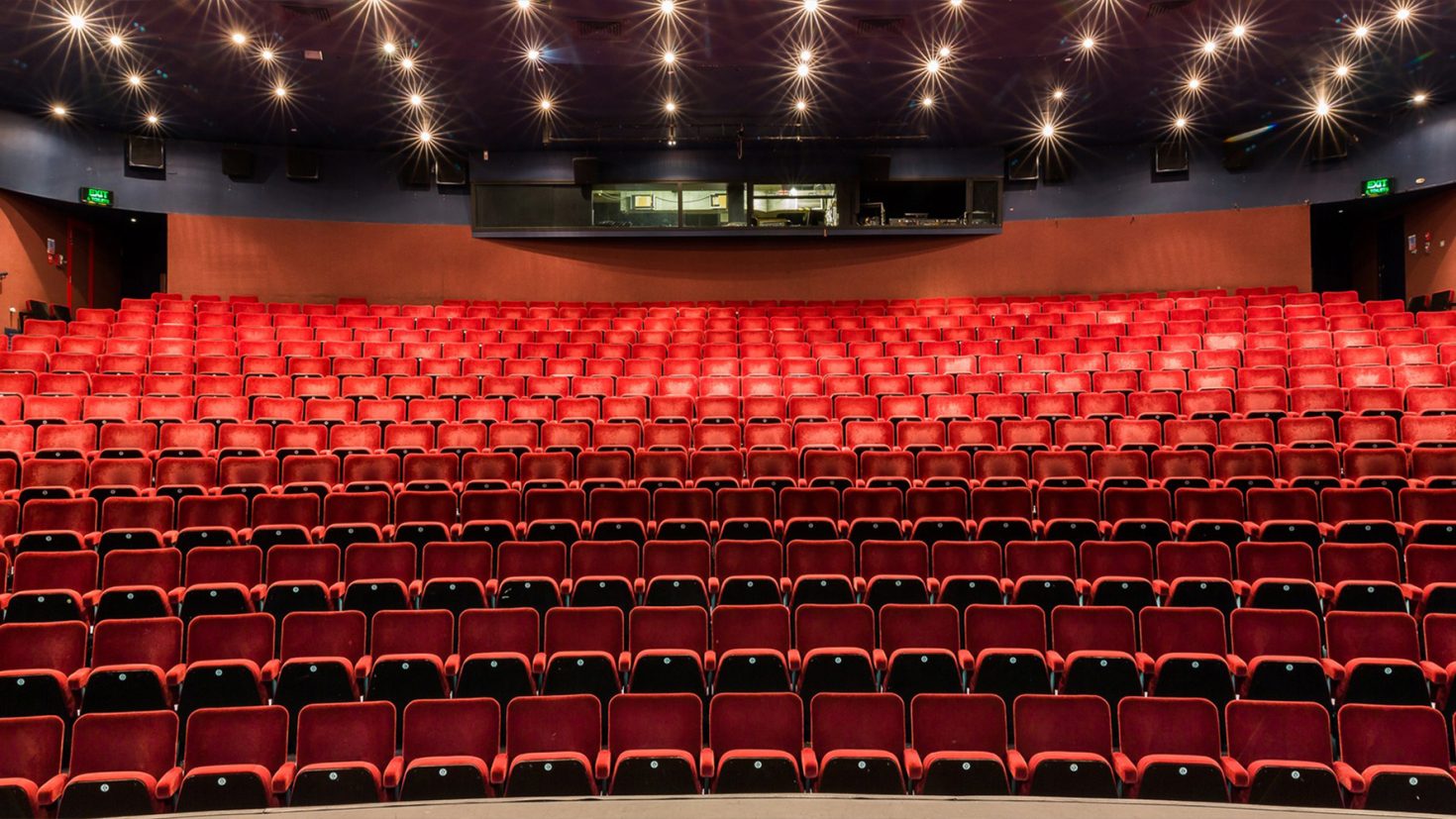 A view of an empty theatre with rows of red seats arranged in a semi-circular layout facing a stage. The ceiling is illuminated with small star-like lights. A control room with a large window is visible at the back of the auditorium.