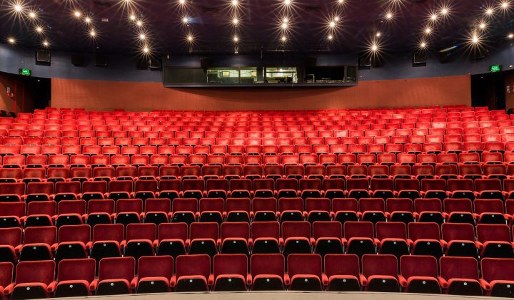 A view of an empty theatre with rows of red seats arranged in a semi-circular layout facing a stage. The ceiling is illuminated with small star-like lights. A control room with a large window is visible at the back of the auditorium.