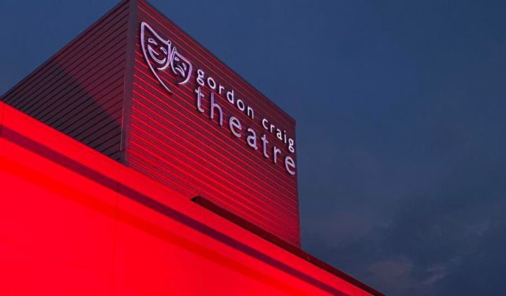 Gordon Craig Theatre building illuminated in red light against a dark evening sky, with its name and a theatre mask logo prominently displayed.