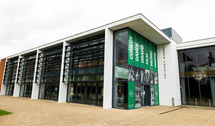 A modern theatre building with large glass windows, labelled "Grove Theatre." The exterior features banners with text "COMEDY," "DRAMA," "DANCE," "FILM," and "MUSIC." The entrance has multiple glass doors and is surrounded by a paved walkway with green grass nearby.