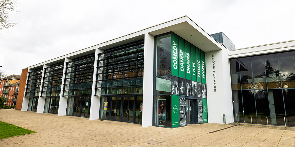 A modern theatre building with large glass windows, labelled "Grove Theatre." The exterior features banners with text "COMEDY," "DRAMA," "DANCE," "FILM," and "MUSIC." The entrance has multiple glass doors and is surrounded by a paved walkway with green grass nearby.