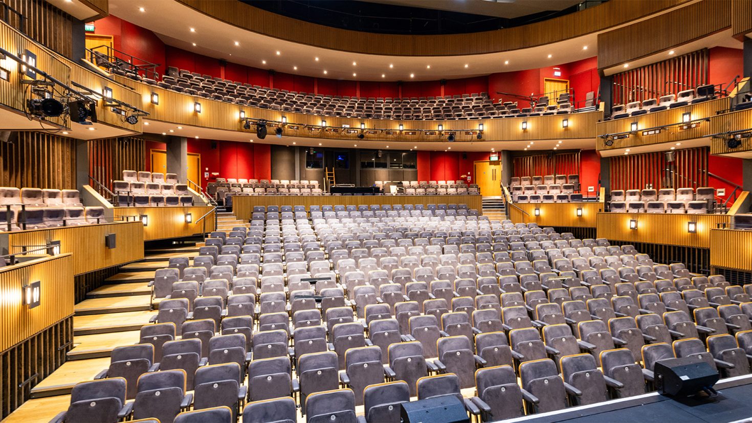 A modern theatre interior with three levels of seating—main floor, mezzanine, and balcony. The seats are upholstered in grey fabric and arranged in rows. The space features wooden accents, red walls, and warm lighting, with a focused spotlight on the stage.