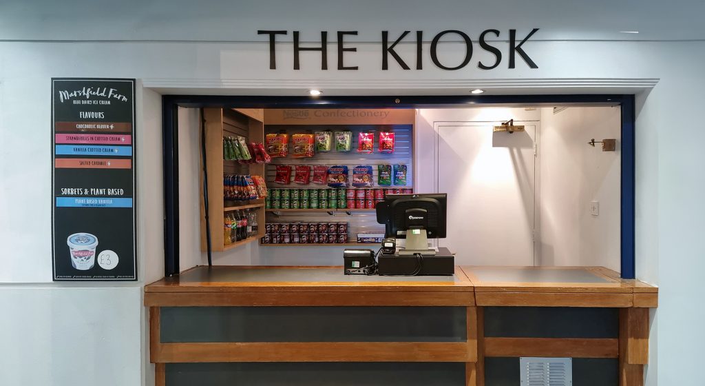 A small kiosk with a wooden counter and a sign overhead reading "THE KIOSK." Behind the counter is a shelf stocked with various snacks and confectioneries. A cash register is placed on the counter, and a menu board is displayed on the left side.