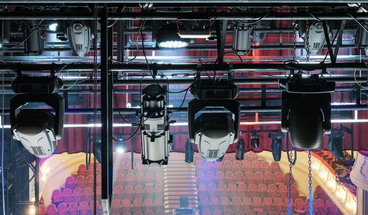 A view from the stage looking out towards an empty auditorium, showing multiple stage lights and equipment hanging from overhead rigs. Rows of red theatre seats and a balcony are visible in the background, as well as the steps leading up to the back of the theatre.