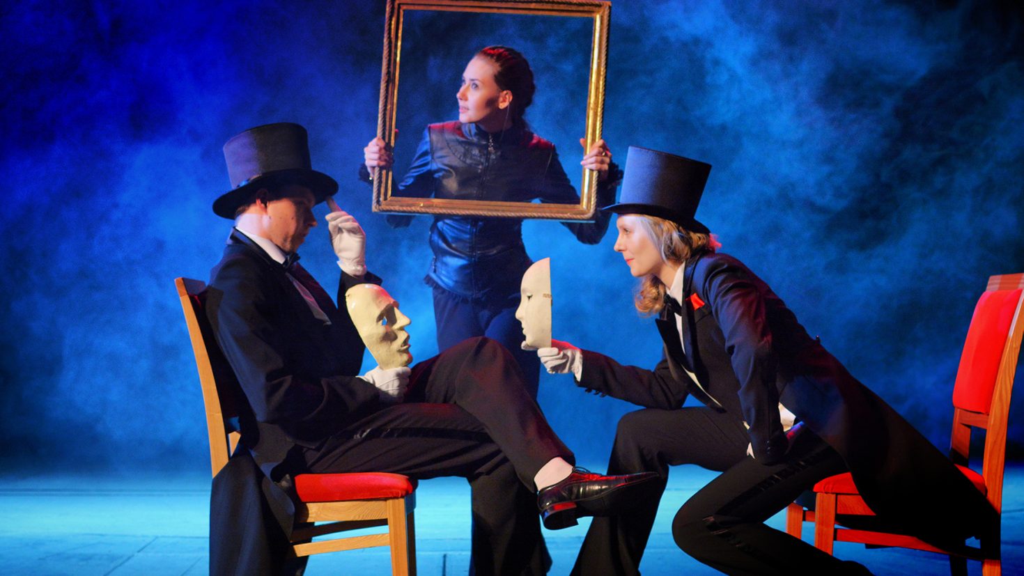 Three performers on stage, shrouded in blue smoke. Two wear black suits, top hats, and white gloves, each holding a mask, sitting on chairs facing each other. A third performer stands behind them, holding a large frame, creating the illusion of a portrait.