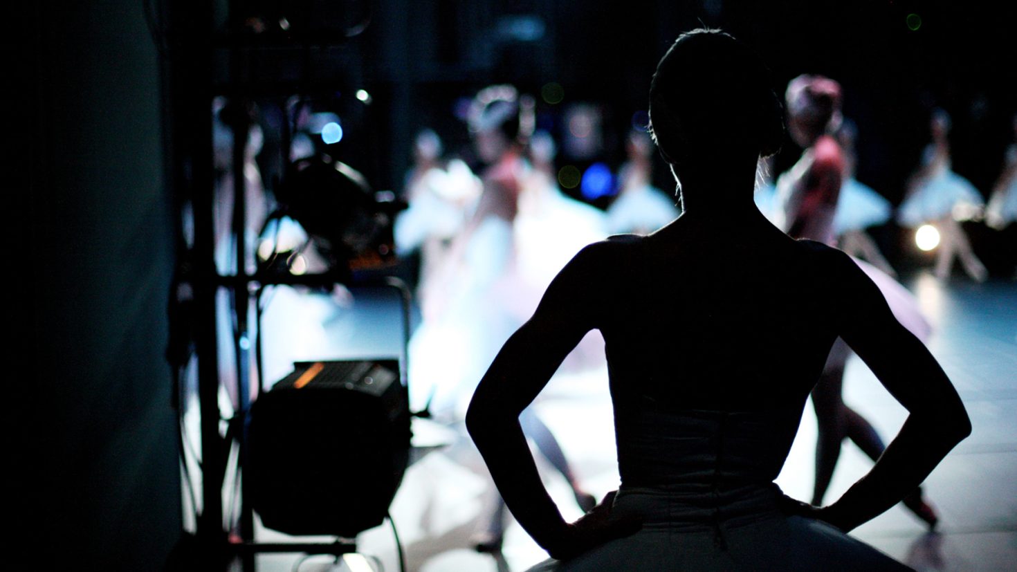 A silhouetted ballet dancer stands backstage with hands on hips, watching fellow dancers perform on stage. The stage lighting highlights the dancers in tutus, creating a vibrant but blurred background against the darkened backstage area.