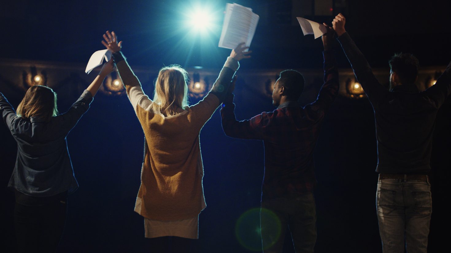 Four actors holding scripts in their hands stand on a stage with their backs to the camera, arms raised in celebration. The stage is lit with a bright spotlight behind them, and the audience seats are dimly visible in the background.