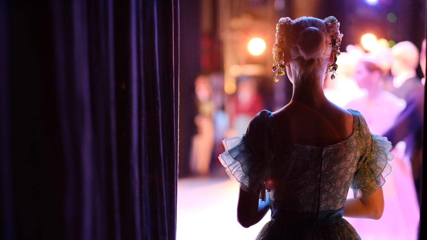 A ballet dancer in an elegant costume with floral hair accessories stands backstage, gazing towards the illuminated stage where other dancers perform. The curtains frame her silhouette, capturing a moment of anticipation and grace.