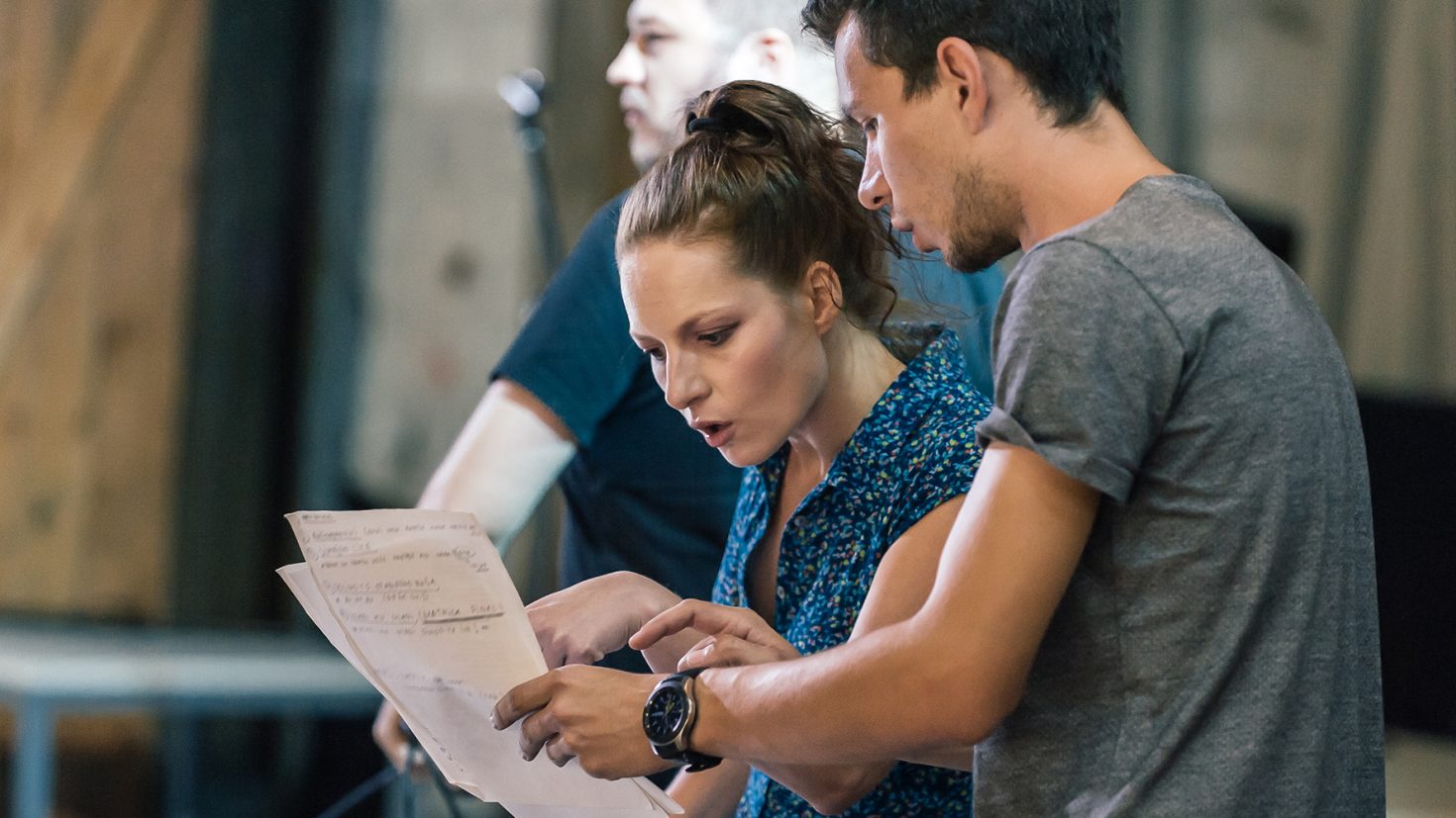 A woman and a man stand closely together, intently discussing a sheet of paper they are holding. The woman, with her hair tied back, appears to be explaining something, while the man, wearing glasses, is focused on the document. Another person is blurred in the background.