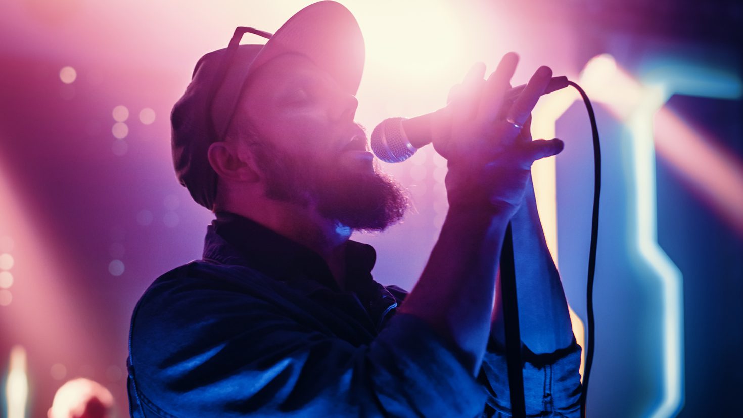 A musician with a beard and cap sings passionately into a microphone under colourful stage lights. He holds the microphone stand with both hands, and the background is illuminated with a vibrant mix of purple, pink, and blue hues, creating a dynamic atmosphere.