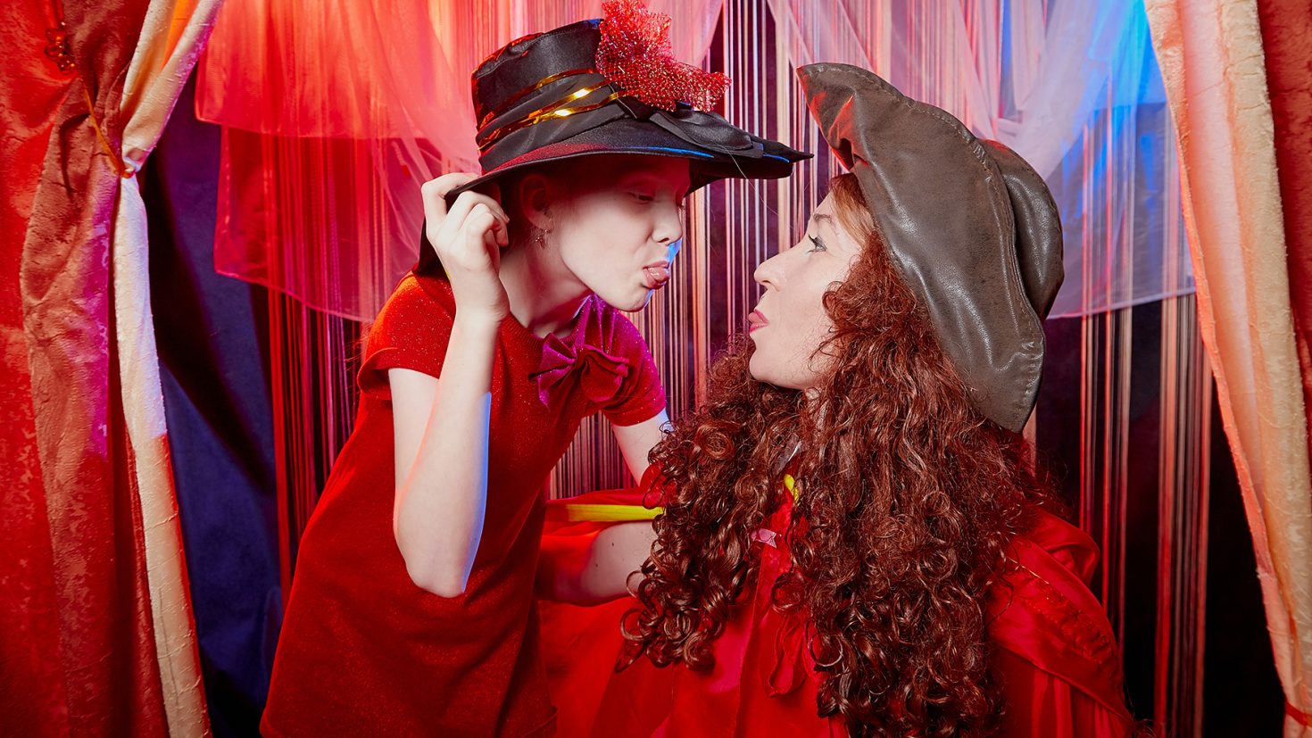 A child and an adult wearing witch hats face each other in a playful manner. The child, dressed in a red outfit, sticks out their tongue while the adult, also in red, leans forward. They are surrounded by colourful drapes, creating a whimsical scene.