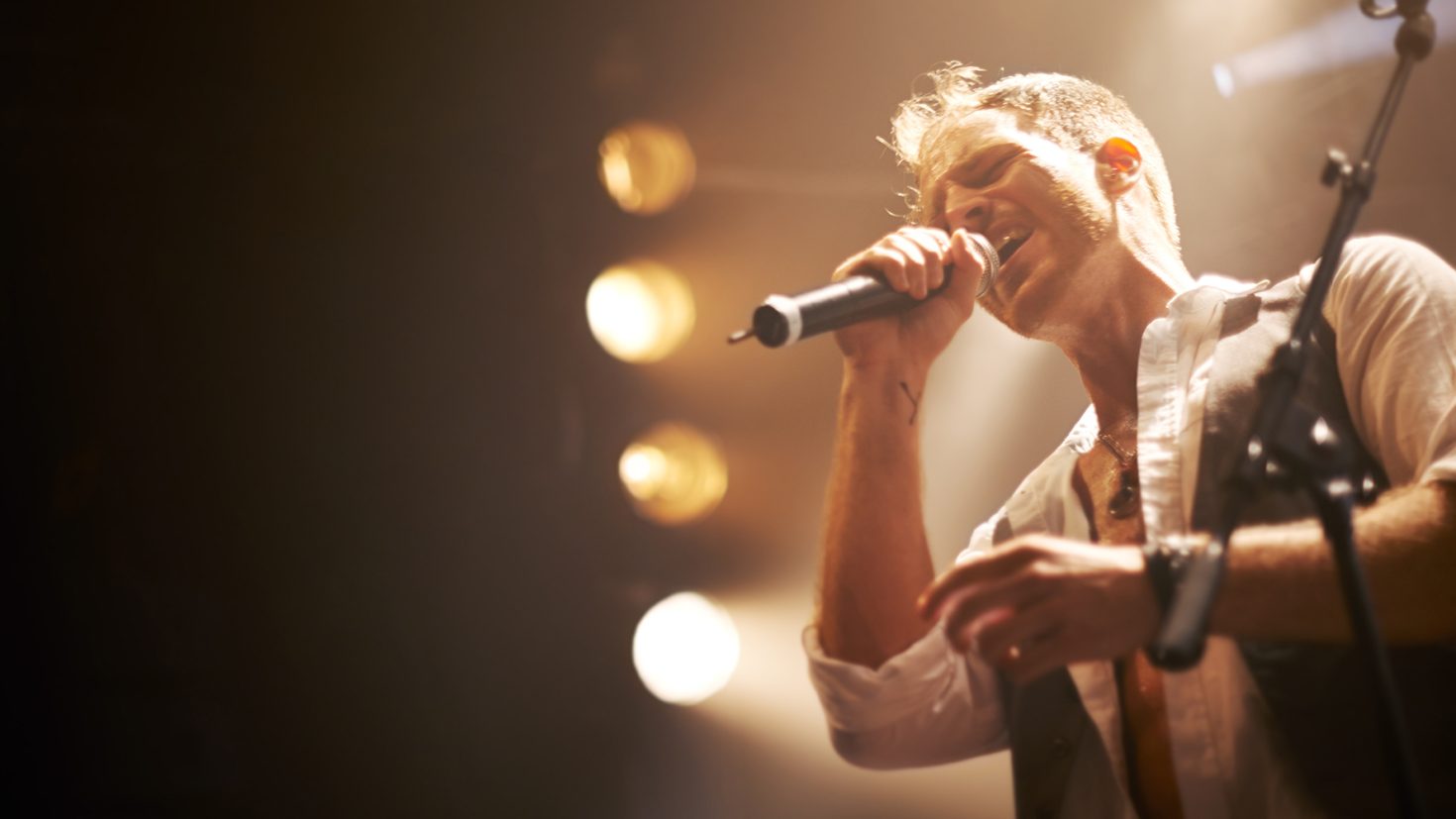 A male singer passionately performs on stage, holding a microphone close to his mouth. The stage lights cast a warm glow around him, emphasizing his expression. He is wearing a white shirt and a vest, and the background is slightly blurred with several bright spotlights.