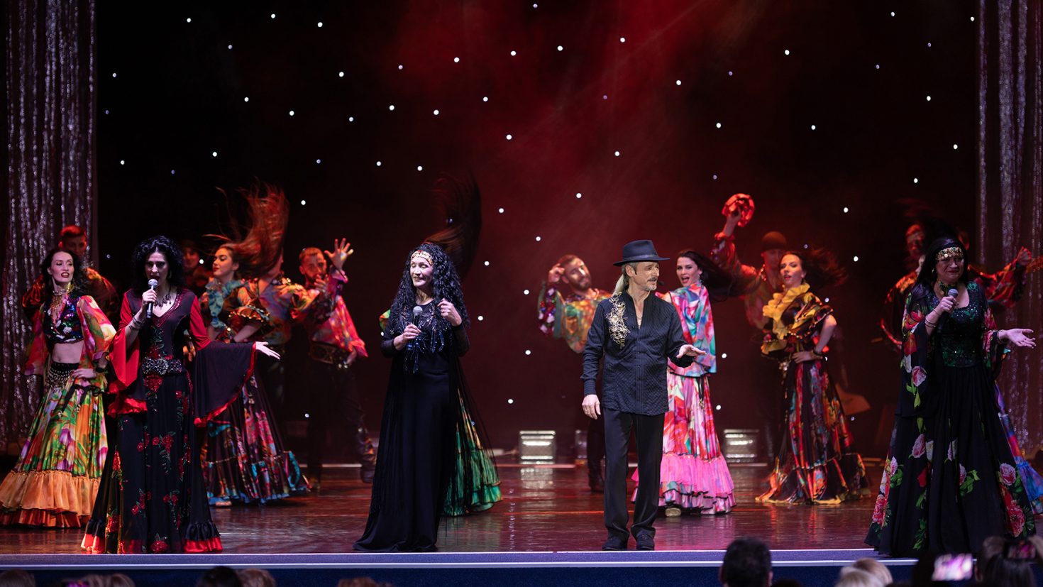 A vibrant stage performance featuring dancers in colourful, traditional costumes. Three performers stand at the front, singing, with one man in a black hat. Sparkling, star-like lights adorn the dark backdrop, and the audience is visible at the bottom of the image.