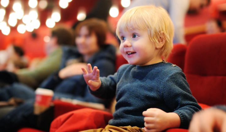 A young child with blonde hair sits in a red theatre seat, smiling excitedly and gesturing with one hand. The background shows several adults seated, also watching the event. The dim lighting and bokeh effect indicate a lively performance or movie screening.