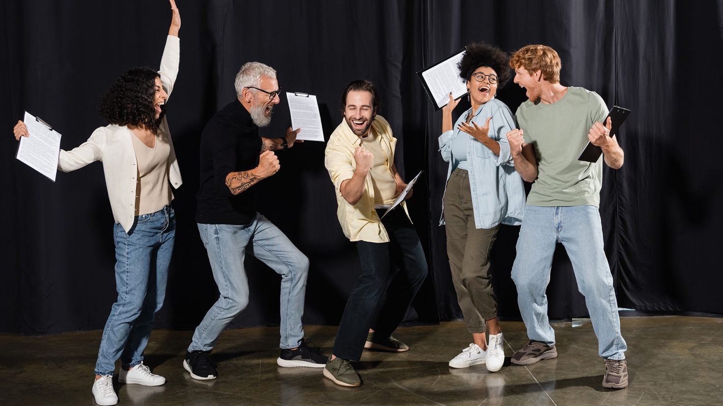 A group of five people stands on a stage with black curtains, each holding scripts or tablets. They appear joyful, raising their arms and waving their hands as if celebrating or cheering. They are dressed casually, in jeans and various tops.