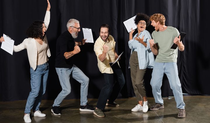 A group of five people stands on a stage with black curtains, each holding scripts or tablets. They appear joyful, raising their arms and waving their hands as if celebrating or cheering. They are dressed casually, in jeans and various tops.