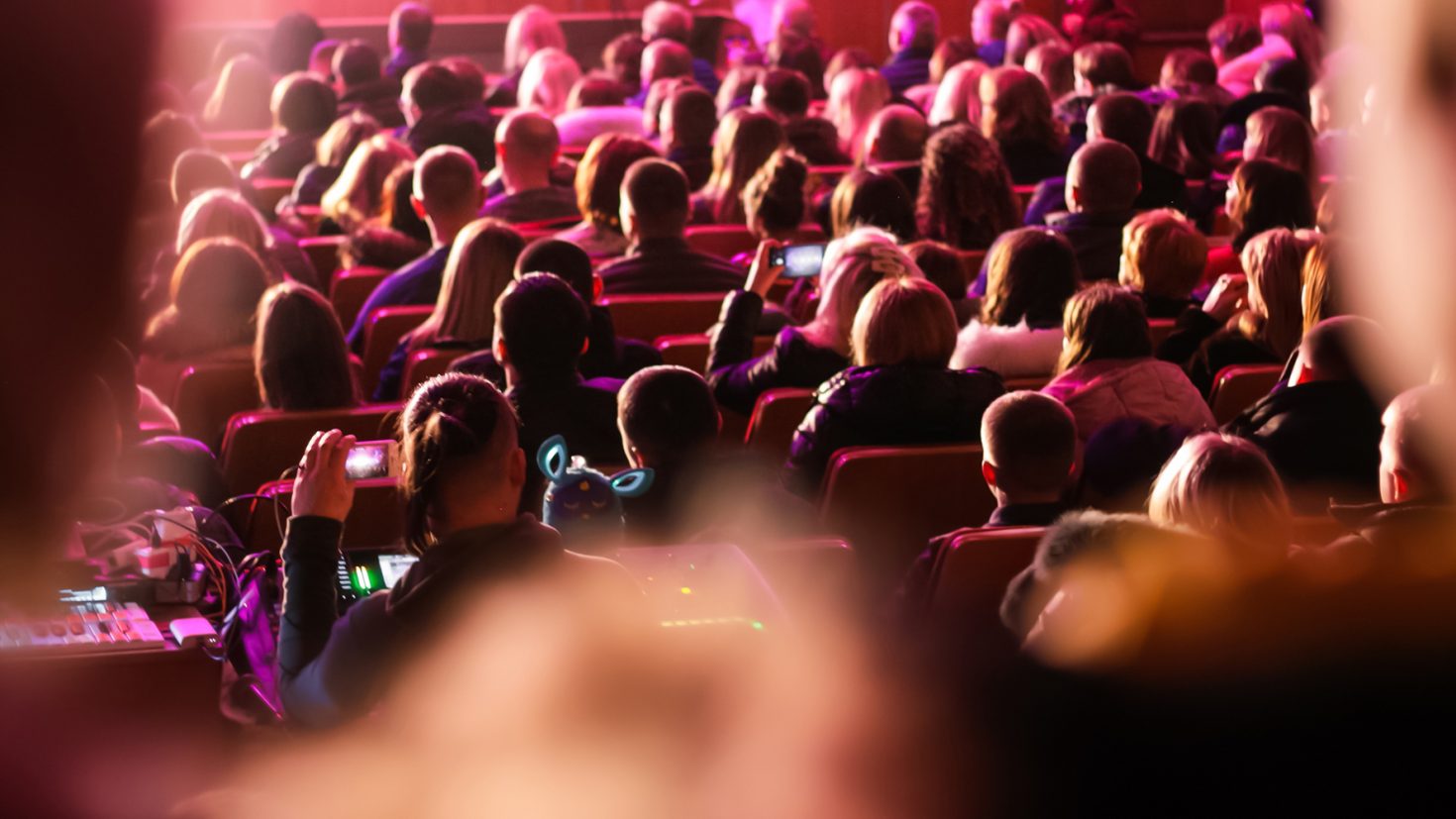 A crowded auditorium filled with people, seen from the back, mostly facing forward. Some attendees are capturing the event with their smartphones. The scene is lit with vibrant pink and red lights, creating a warm and lively atmosphere.