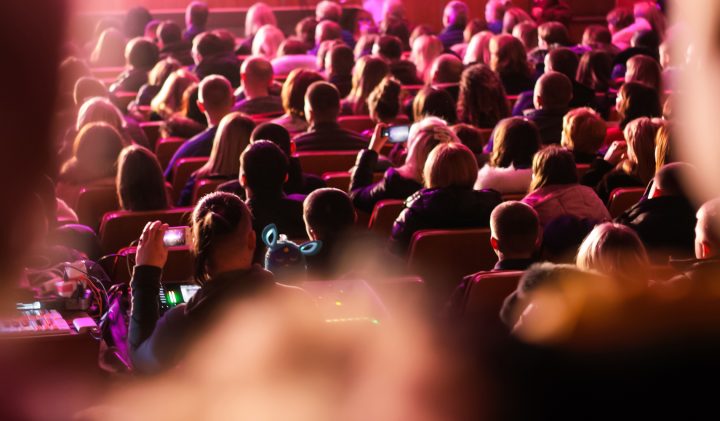 A crowded auditorium filled with people, seen from the back, mostly facing forward. Some attendees are capturing the event with their smartphones. The scene is lit with vibrant pink and red lights, creating a warm and lively atmosphere.